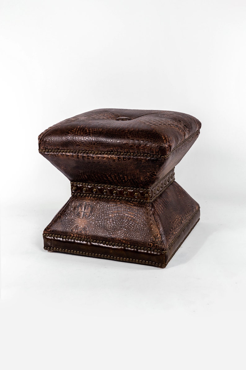 K&ouml;nnte beinhalten: Ein brauner Lederhocker mit quadratischer Oberseite und einem dekorativen Sockel. Der Hocker hat ein strukturiertes, krokodil&auml;hnliches Muster und ist mit Metallnieten verziert.