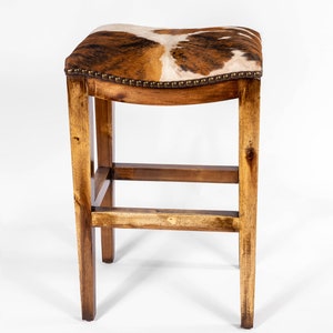 May include: A wooden bar stool with a saddle-shaped seat upholstered in brown and white cowhide. The stool has a simple, rustic design with a rectangular frame and a single crossbar.
