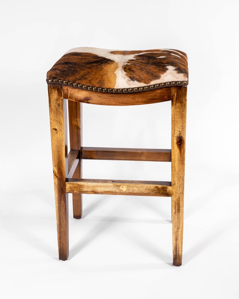May include: A wooden bar stool with a saddle-shaped seat upholstered in brown and white cowhide. The stool has a simple, rustic design with a rectangular frame and a single crossbar.