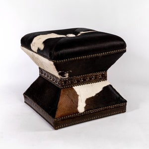 K&ouml;nnte beinhalten: Ein quadratischer Hocker mit einem Muster aus schwarzem, wei&szlig;em und braunem Kuhfell. Der Hocker hat eine gesteppte Oberseite und einen dekorativen Metallrand an den Kanten.