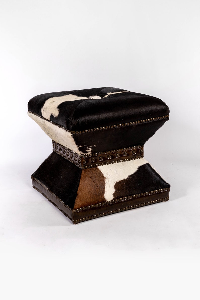 K&ouml;nnte beinhalten: Ein quadratischer Hocker mit einem Muster aus schwarzem, wei&szlig;em und braunem Kuhfell. Der Hocker hat eine gesteppte Oberseite und einen dekorativen Metallrand an den Kanten.