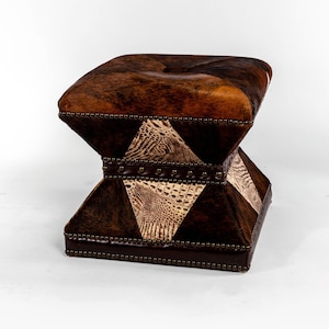 May include: A brown and tan leather ottoman with a diamond pattern and studded trim. The ottoman is made of cowhide and has a unique, rustic look.