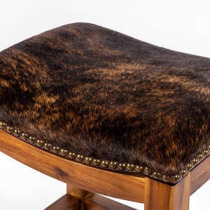 May include: A brown cowhide bar stool with a wooden frame and a rounded seat. The seat is upholstered in brown cowhide with a dark brown and light brown pattern.