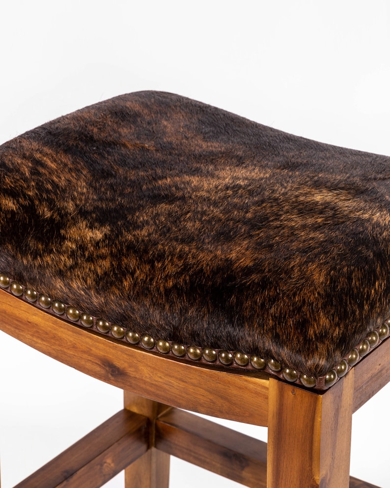 May include: A brown cowhide bar stool with a wooden frame and a rounded seat. The seat is upholstered in brown cowhide with a dark brown and light brown pattern.