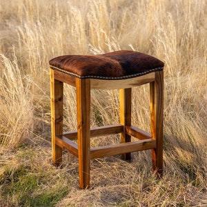 May include: A brown leather bar stool with a wooden frame. The stool is sitting on a field of tall grass.