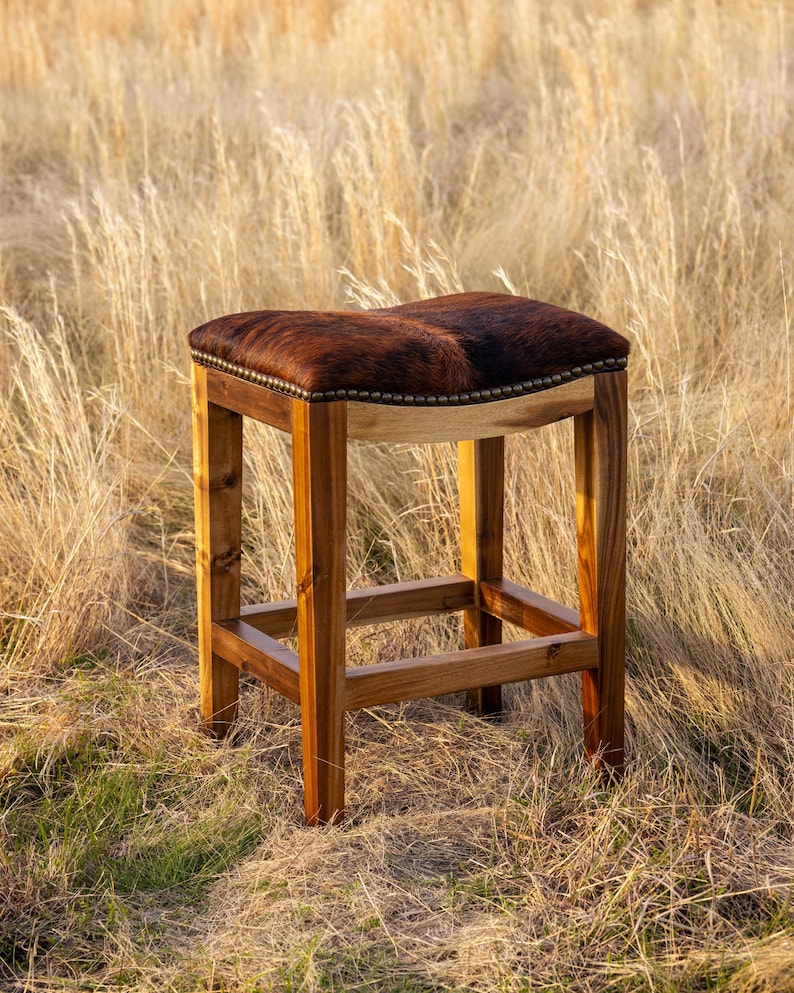 May include: A brown leather bar stool with a wooden frame. The stool is sitting on a field of tall grass.