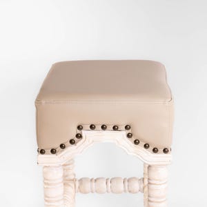May include: White wooden stool with a beige upholstered seat and decorative nailhead trim. The stool has a carved design on the legs and a curved back.
