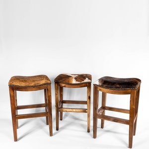 May include: Three wooden bar stools with brown cowhide seats. The stools have a simple design with a single crossbar and four legs.