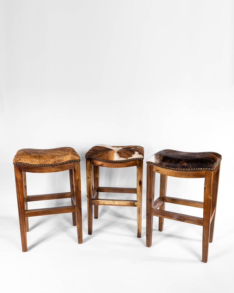 May include: Three wooden bar stools with brown cowhide seats. The stools have a simple design with a single crossbar and four legs.