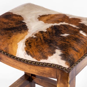 May include: A wooden bar stool with a brown cowhide seat. The seat is upholstered with a brown, white, and black cowhide pattern. The stool has a rounded top and a simple, sturdy design.