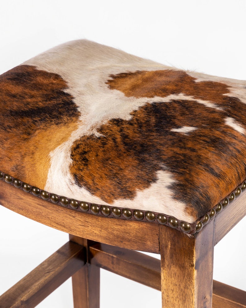 May include: A wooden bar stool with a brown cowhide seat. The seat is upholstered with a brown, white, and black cowhide pattern. The stool has a rounded top and a simple, sturdy design.