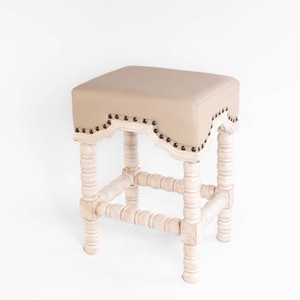 May include: A white wooden stool with a beige upholstered seat. The seat has a decorative scalloped edge with metal studs. The legs are turned and have a distressed white finish.
