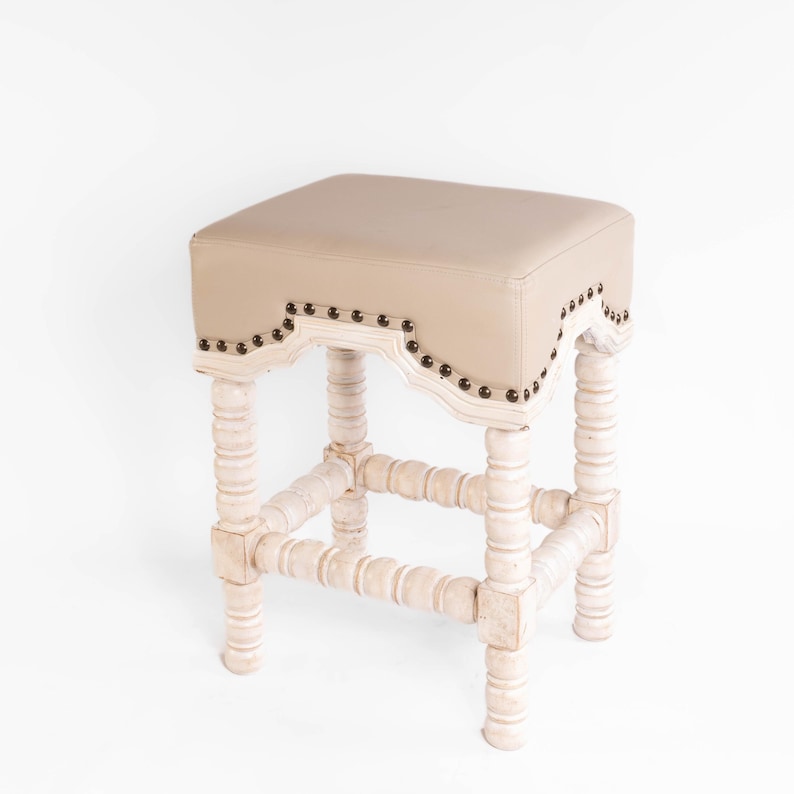 May include: A white wooden stool with a beige upholstered seat. The seat has a decorative scalloped edge with metal studs. The legs are turned and have a distressed white finish.