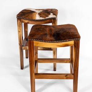 May include: Two wooden bar stools with brown cowhide seats. The stools have a simple design with a rectangular base and a curved seat.