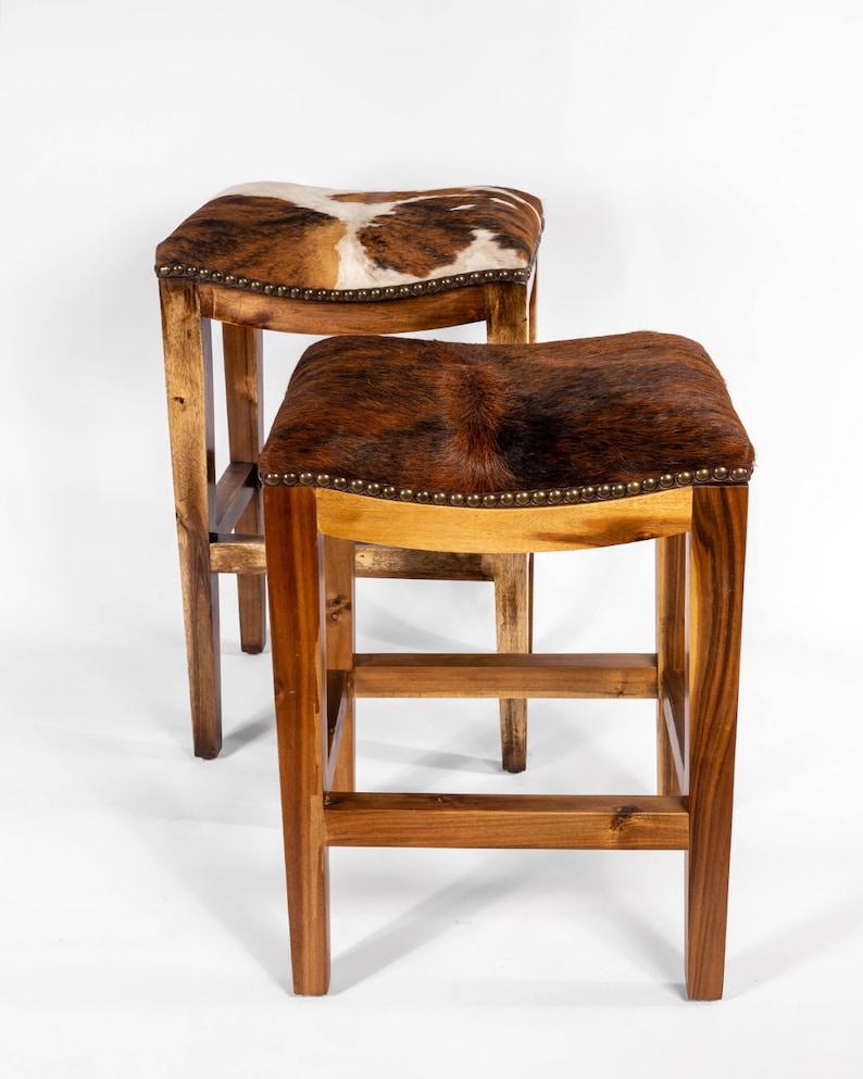 May include: Two wooden bar stools with brown cowhide seats. The stools have a simple design with a rectangular base and a curved seat.