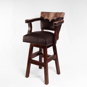 May include: A brown leather bar stool with a high back and armrests. The backrest features a decorative patterned leather insert. The stool has a dark wooden frame and a padded seat, with brass nailhead trim.
