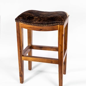 May include: A brown wooden bar stool with a saddle-shaped seat upholstered in brown and white cowhide. The stool has a simple, rustic design with a single crossbar for support.