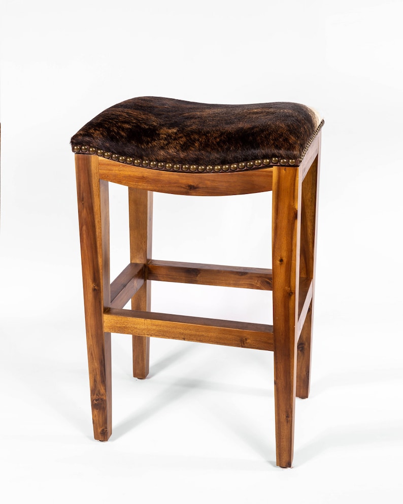 May include: A brown wooden bar stool with a saddle-shaped seat upholstered in brown and white cowhide. The stool has a simple, rustic design with a single crossbar for support.