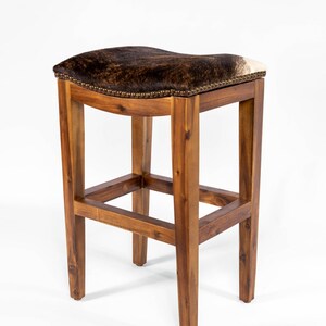 May include: A brown wooden bar stool with a saddle-shaped seat upholstered in brown and white cowhide. The stool has a simple, rustic design with a rectangular frame and four legs.