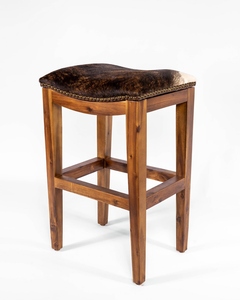 May include: A brown wooden bar stool with a saddle-shaped seat upholstered in brown and white cowhide. The stool has a simple, rustic design with a rectangular frame and four legs.