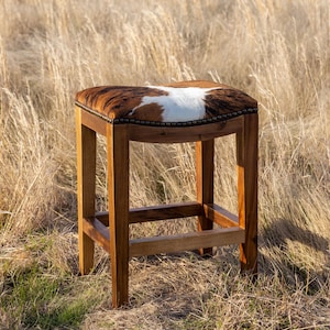 May include: A brown wooden bar stool with a cowhide seat. The stool is in a field of tall grass.