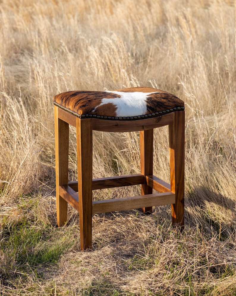 May include: A brown wooden bar stool with a cowhide seat. The stool is in a field of tall grass.
