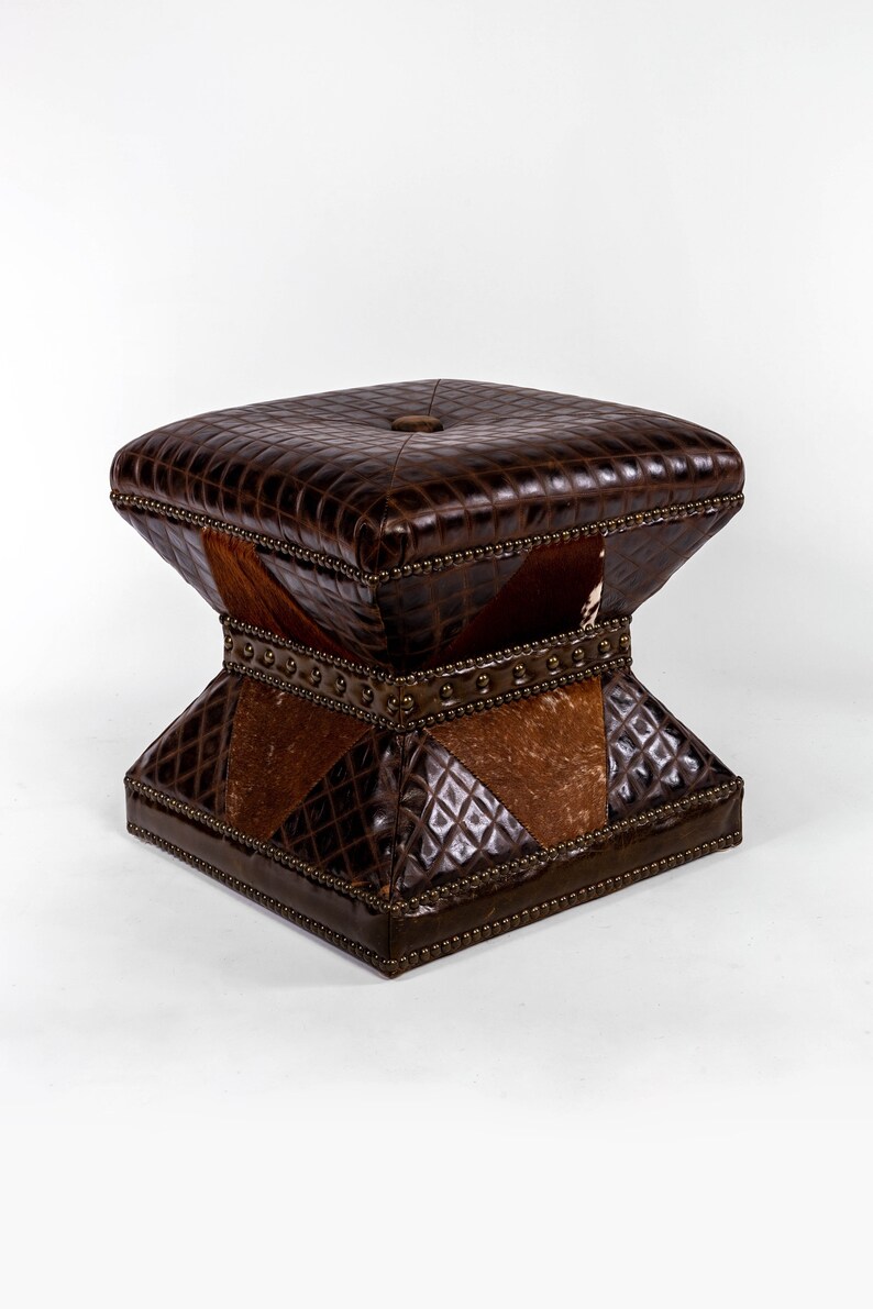 K&ouml;nnte beinhalten: Ein brauner Lederhocker mit quadratischer Oberseite und einem einzigartigen, geometrischen Design. Der Hocker besteht aus verschiedenen Ledersorten, darunter ein braunes Leder mit Kroko-Muster und ein braunes und wei&szlig;es Kuhfellmuster.
