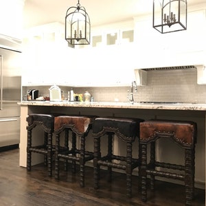 May include: Four brown leather bar stools with studded details and a dark wood frame. The stools are arranged in a row in front of a kitchen island with a granite countertop.