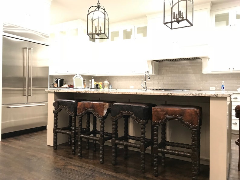 May include: Four brown leather bar stools with studded details and a dark wood frame. The stools are arranged in a row in front of a kitchen island with a granite countertop.