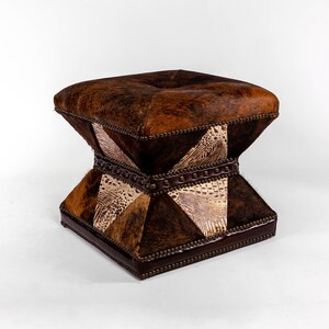 K&ouml;nnte beinhalten: Ein quadratischer Hocker mit einem braunen, wei&szlig;en und schwarzen Patchwork-Design. Der Hocker ist mit Leder bezogen und hat eine gesteppte Oberseite.