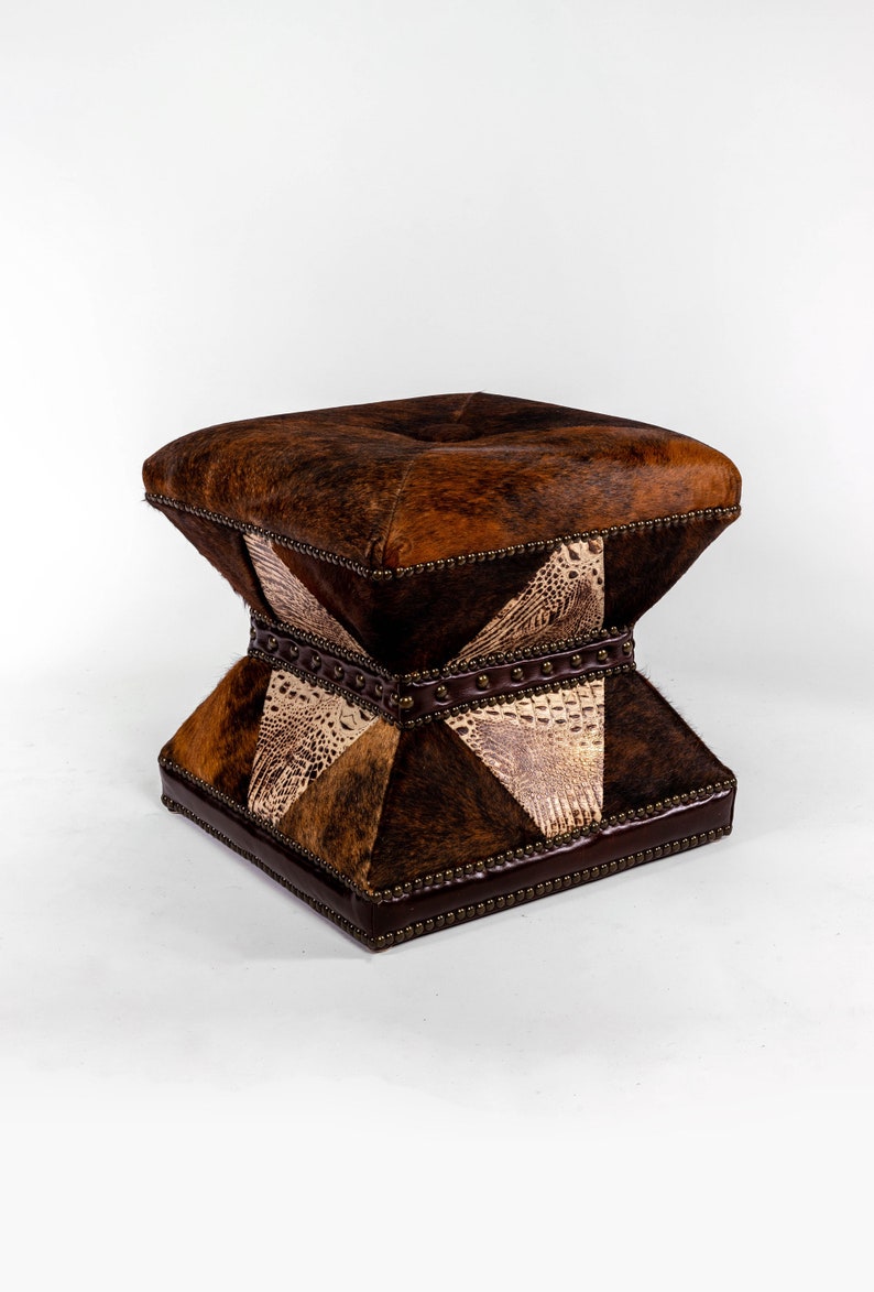 K&ouml;nnte beinhalten: Ein quadratischer Hocker mit einem braunen, wei&szlig;en und schwarzen Patchwork-Design. Der Hocker ist mit Leder bezogen und hat eine gesteppte Oberseite.