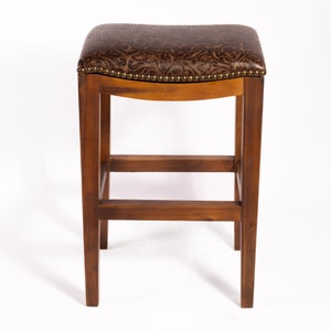 May include: A brown wooden bar stool with a padded seat upholstered in brown faux leather with a decorative pattern. The seat has a rounded edge and is accented with metal studs.