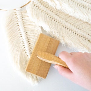 May include: A close-up of a wooden brush with a natural finish and a wooden handle. The brush is being used to smooth out a white macrame wall hanging.