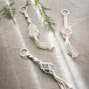 Op de afbeelding: Drie macramé sleutelhangers met verschillende gekleurde kristallen. Elke sleutelhanger is gemaakt van wit touw en heeft een zilveren sleutelring. De kristallen zijn helder, roze en groen.