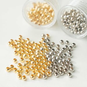 May include: A pile of gold and silver beads, scattered on a white surface. The beads are small and round, and they are arranged in a random pattern.