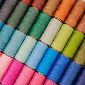 Puede incluir: Una colección de coloridas bobinas de hilo, dispuestas en diagonal. Los hilos son de varios tonos, incluyendo azul, verde, rosa, naranja y marrón. Las bobinas son de madera y el hilo está bien enrollado.