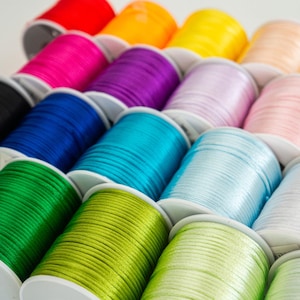 Può includere: Una collezione di bobine di cordoncino di raso colorato, ciascuna avvolta attorno a una bobina di plastica bianca. I cordoncini sono disposti a semicerchio, con il cordoncino rosso in alto e il cordoncino verde in basso.