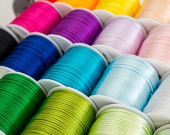 May include: A collection of spools of colorful satin cord, each wound around a white plastic bobbin. The cords are arranged in a semi-circle, with the red cord at the top and the green cord at the bottom.