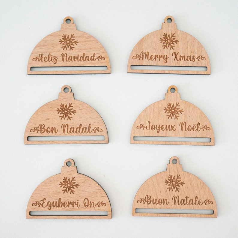 May include: Five wooden Christmas ornament gift tags with a snowflake design and a slot for a gift card. The tags read "Feliz Navidad", "Merry Xmas", "Bon Nadal", "Joyeux Noel", and "Buon Natale".