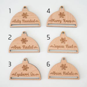 May include: Six wooden ornaments shaped like Christmas tree ornaments with a snowflake design and a slot for a gift tag. The ornaments are labeled with the numbers 1 through 6 and have the following text: "Feliz Navidad", "Bon Nadal", "Eguberri On", "Merry Xmas", "Joyeux No&euml;l", and "Buon Natale".