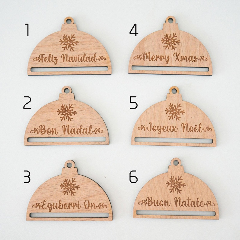 May include: Six wooden ornaments shaped like Christmas tree ornaments with a snowflake design and a slot for a gift tag. The ornaments are labeled with the numbers 1 through 6 and have the following text: "Feliz Navidad", "Bon Nadal", "Eguberri On", "Merry Xmas", "Joyeux No&euml;l", and "Buon Natale".