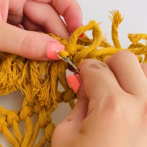 Könnte beinhalten: Nahaufnahme von Händen, die einen Häkelhaken verwenden, um Fäden aus gelbem Garn zu verweben. Das Garn wird zu einer Makramee-Wandbehang verarbeitet.