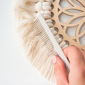 May include: A close-up of a macrame wall hanging being combed with a light beige comb. The macrame is cream-colored with a wooden decorative element. The comb is made of a light beige material.