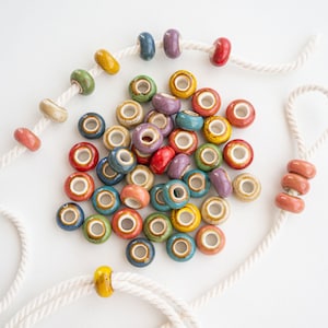 Può includere: Una collezione di perline in ceramica colorate in varie tonalità di blu, verde, giallo, rosso e viola. Le perline sono infilate su un cordoncino bianco e sono perfette per realizzare gioielli o altri oggetti artigianali.