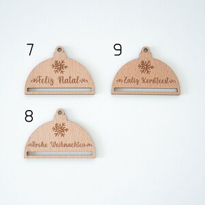 May include: Three wooden Christmas ornaments with a snowflake design and a slot for a gift card. The ornaments read "Feliz Natal", "Zalg Kersfeest", and "Frohe Weihnachten".
