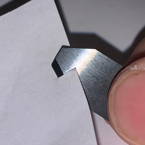 May include: A close-up of a silver paper trimmer blade cutting through a piece of white paper.