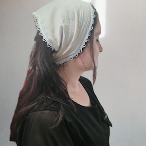 May include: A cream-colored triangular headscarf made of textured fabric, featuring a delicate white lace trim along the edges. The headscarf is tied securely, covering the top and back of the head with a soft, gathered appearance.