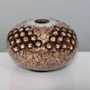 Può includere: Un vaso rotondo in ceramica con esterno maculato marrone e bianco. Il vaso presenta un motivo di cerchi marroni scuri in rilievo. Ha una piccola apertura sulla parte superiore. Il vaso è su una superficie bianca.