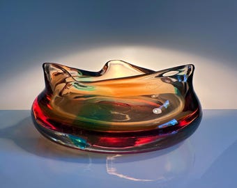 Könnte beinhalten: Eine dekorative Glasschale mit einem einzigartigen, wellenförmigen Rand. Die Schale weist eine Mischung aus Farben auf, darunter Rot, Orange und Grün, wodurch ein lebendiges und künstlerisches Aussehen entsteht. Das Glas hat eine glänzende Oberfläche, die das Licht reflektiert.