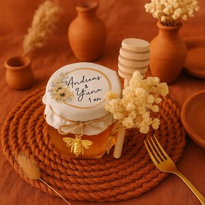 Peut inclure: Un petit pot de miel en verre, surmonté d'un couvercle en tissu avec des motifs floraux et du texte. Un charm en forme d'abeille dorée est suspendu au pot. Un dipper à miel en bois et une fourchette dorée sont également présents.
